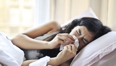 Homöopathie bei Erkältung: zu sehen ist eine junge Frau, die seitlich im Bett liegt und sich die Nase putzt